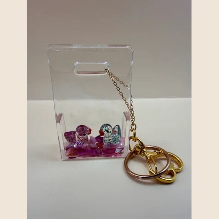 Deze handgemaakte transparante sleutelhanger is een vrolijk en uniek item met twee schattige eendjes en kleine decoratieve steentjes aan de onderkant. Door de heldere uitstraling lijkt het net alsof de eendjes samen in een klein waterwereldje staan. Leuk voor aan je sleutelbos, tas, rugzak of als klein cadeautje voor iemand die dol is op vrolijke, handgemaakte accessoires.